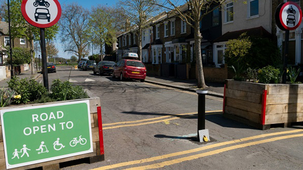 street scene showing parked cars on side of street, yellow lines painted on road, trees, sign prohibiting cars and motorbikes, green sign saying 'Road Open to' and then symbols for pedestrians, skaters, wheelchair users, and cyclists. Two large wooden plant boxes placed on either side of the road.