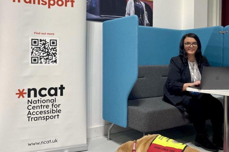 Emma Partlow seated at enclosed desk with assistance dog Luna by her side. To her left is a large standing poster for ncat which reads 'Shaping the future of accessible transport' and has a qr code on it.