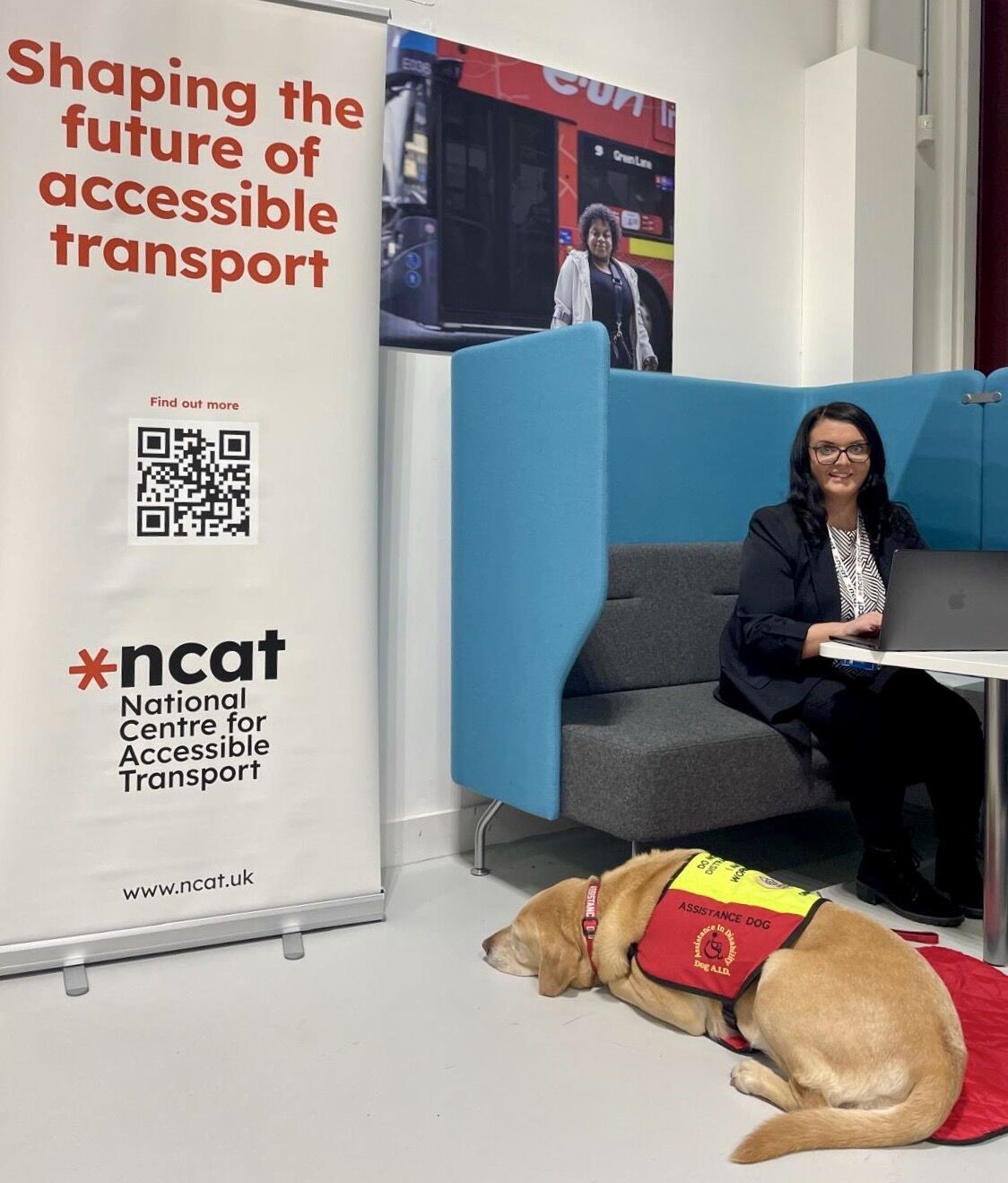 Emma Partlow seated at enclosed desk with assistance dog Luna by her side. To her left is a large standing poster for ncat which reads 'Shaping the future of accessible transport' and has a qr code on it.