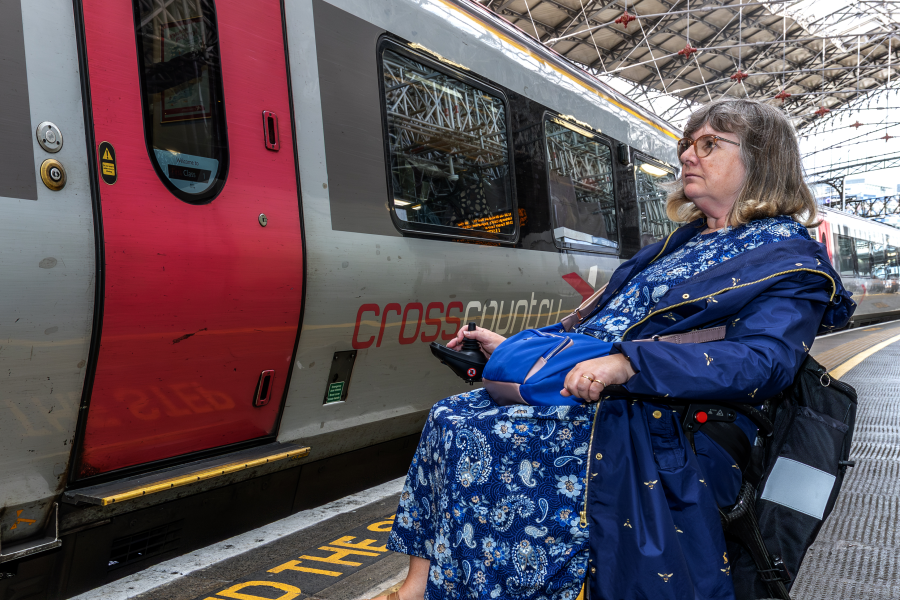 Female wheelchair user in blue dress waits on train platform behind tactile paving. Train, tracks and station to her left in the image.